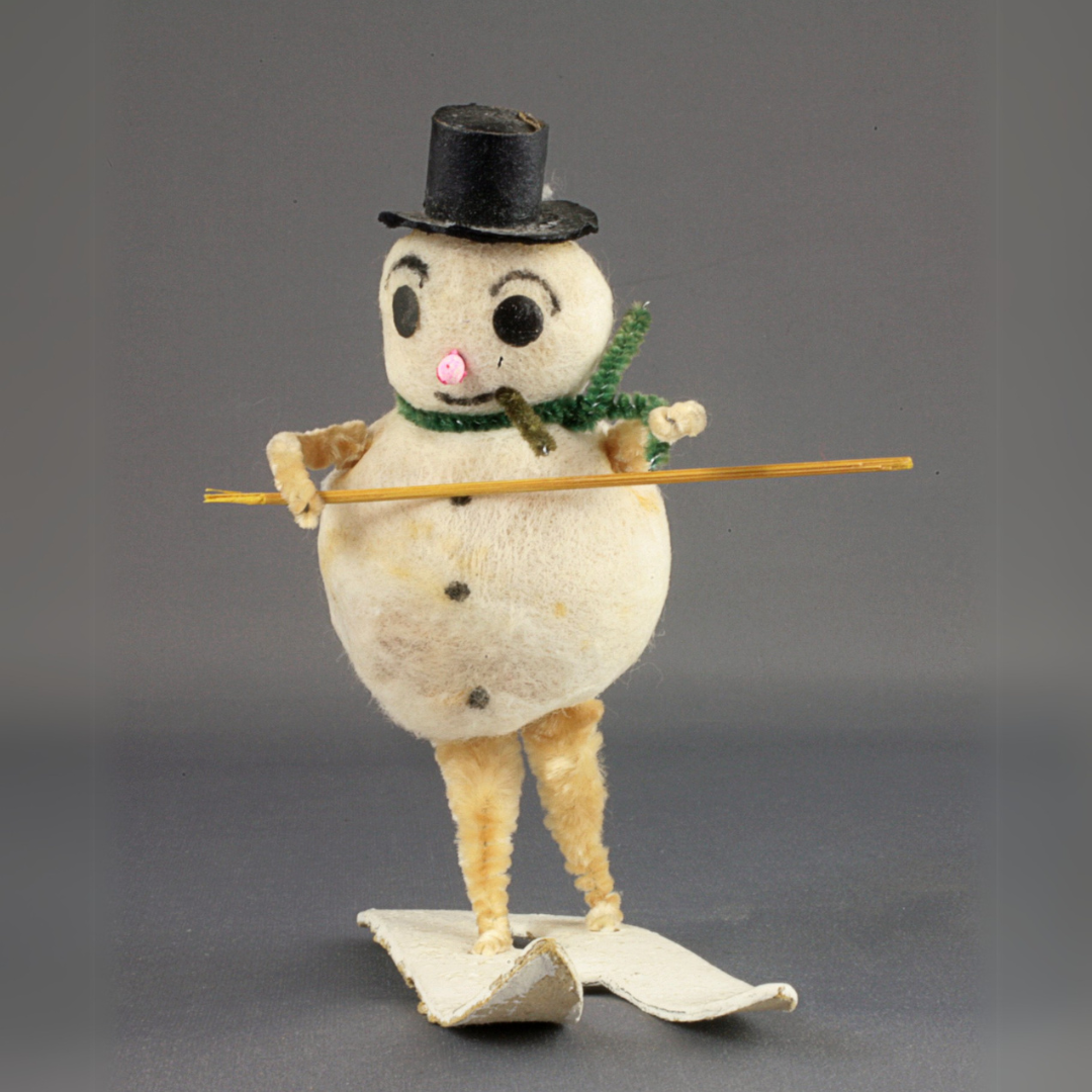 A vintage Frosty the Snowman decoration showing a snowman with legs and arms riding a set of skis while wearing a black top hat and green pipe cleaner scarf.