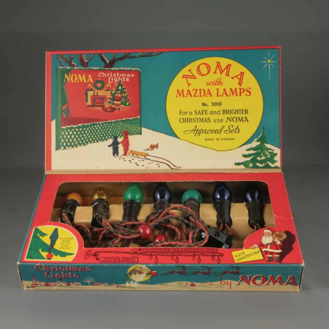 An open vintage cardboard box with Christmas lights arranged inside. The inside of the upper flap shows a winter scene with two children pulling a sled in front of a NOMA Christmas Lights billboard. Text beside the picture reads, "NOMA with Mazda Lamps / For a SAFE and BRIGHTER CHRISTMAS use NOMA / Approved Sets / Made in Canada".