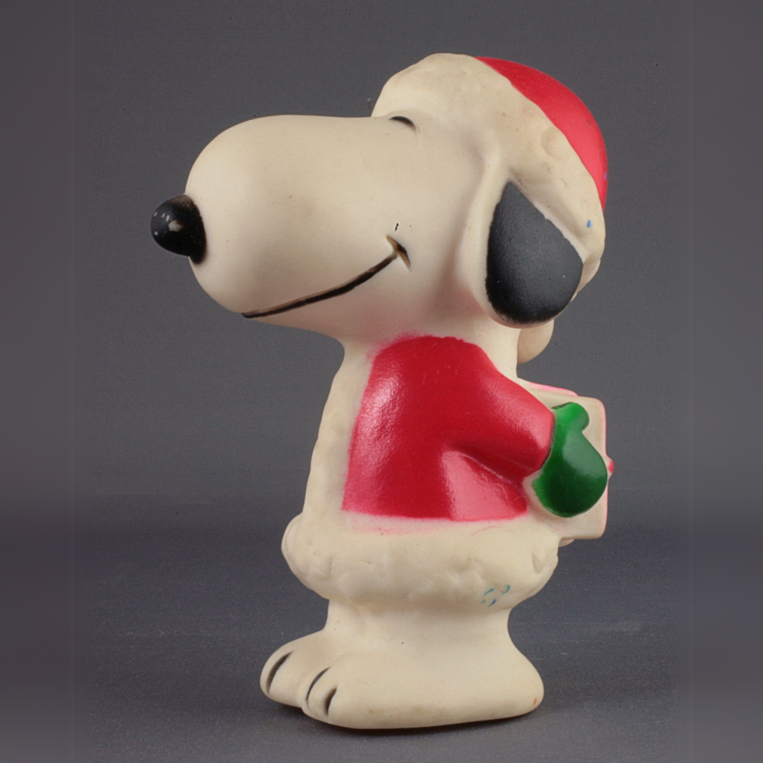 Toy in profile of Snoopy the dog wearing a red and white Santa coat and hat and green mittens, and holding a gift behind his back.