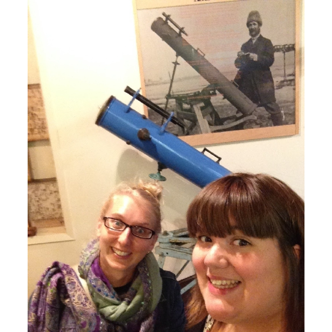 A smiling selfie taken by Cortney of herself and Carolyn standing in front of a blue telescope. On the wall behind the telescope is a black and white photo of Percy Criddle with the same telescope.