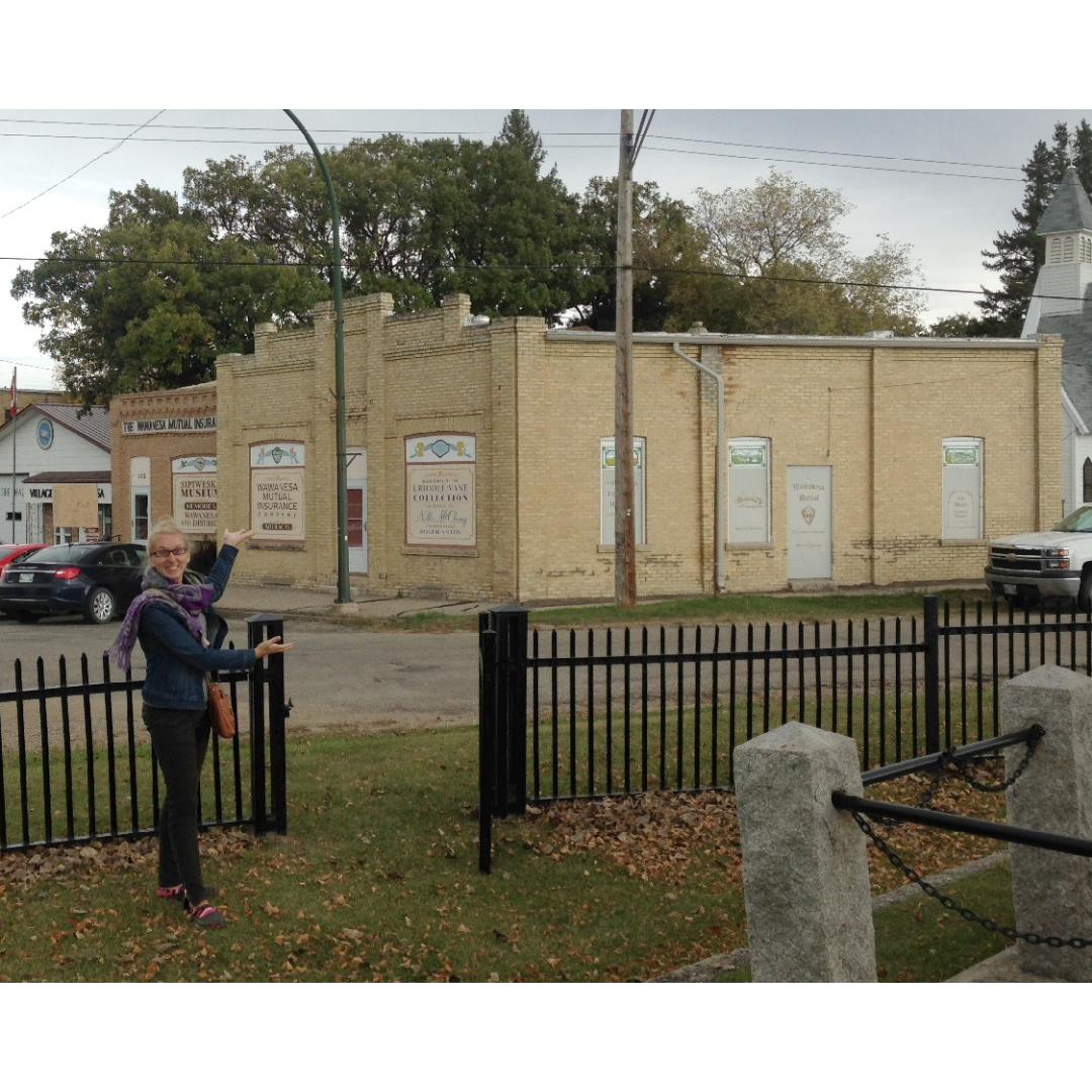 Carolyn stand in a fenced yard gesturing towards a stone building across the street - the museum.