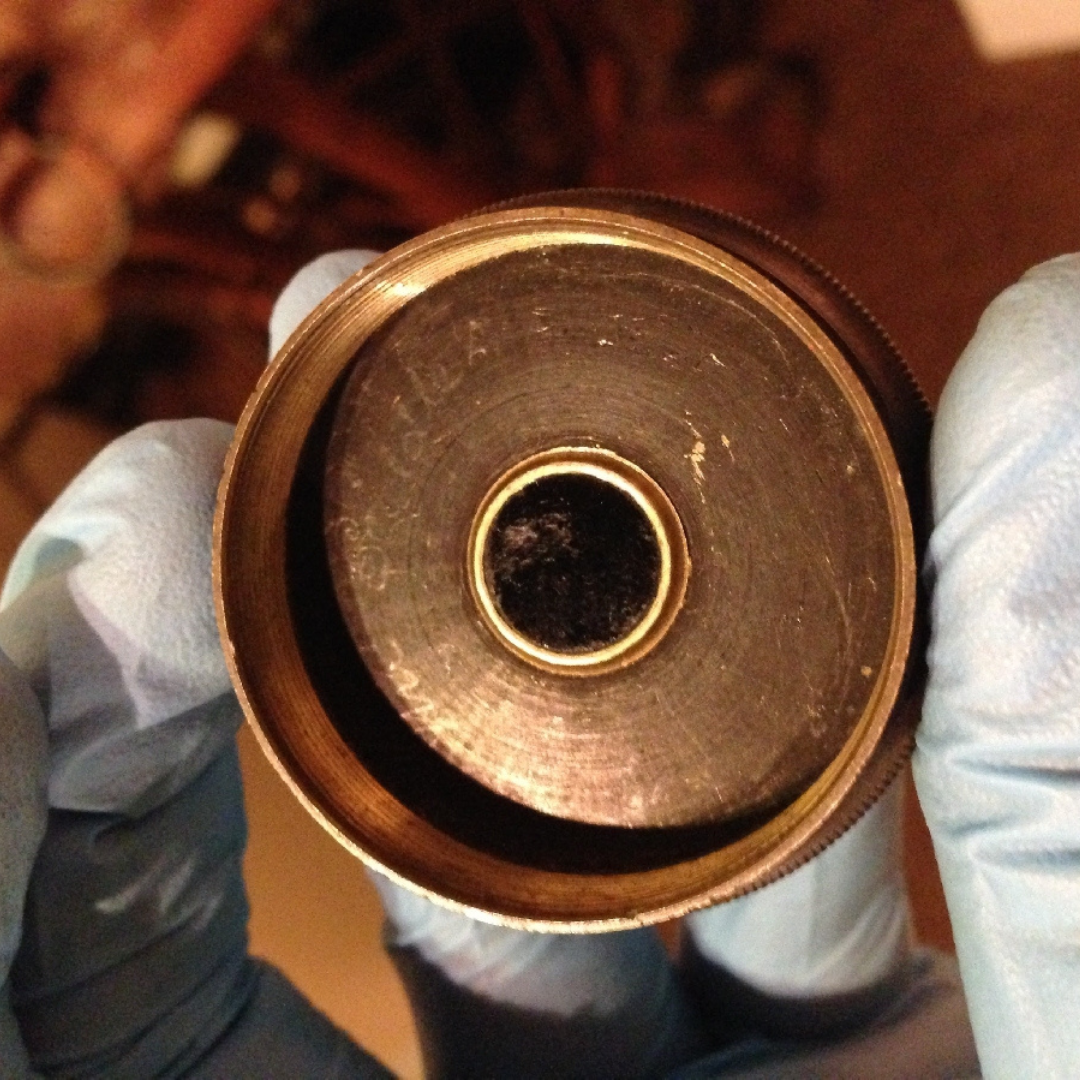 A blu-gloved hand holding up a copper-coloured lens cap with "Percy Criddle" engraved into it by hand.