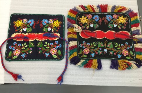 Two intricately beaded square dog blankets. The one on the right has a colourful fringe around the edges, and the one of the left does not, instead having two longer braids as ties. The pattern on the body of each piece is nearly identical.