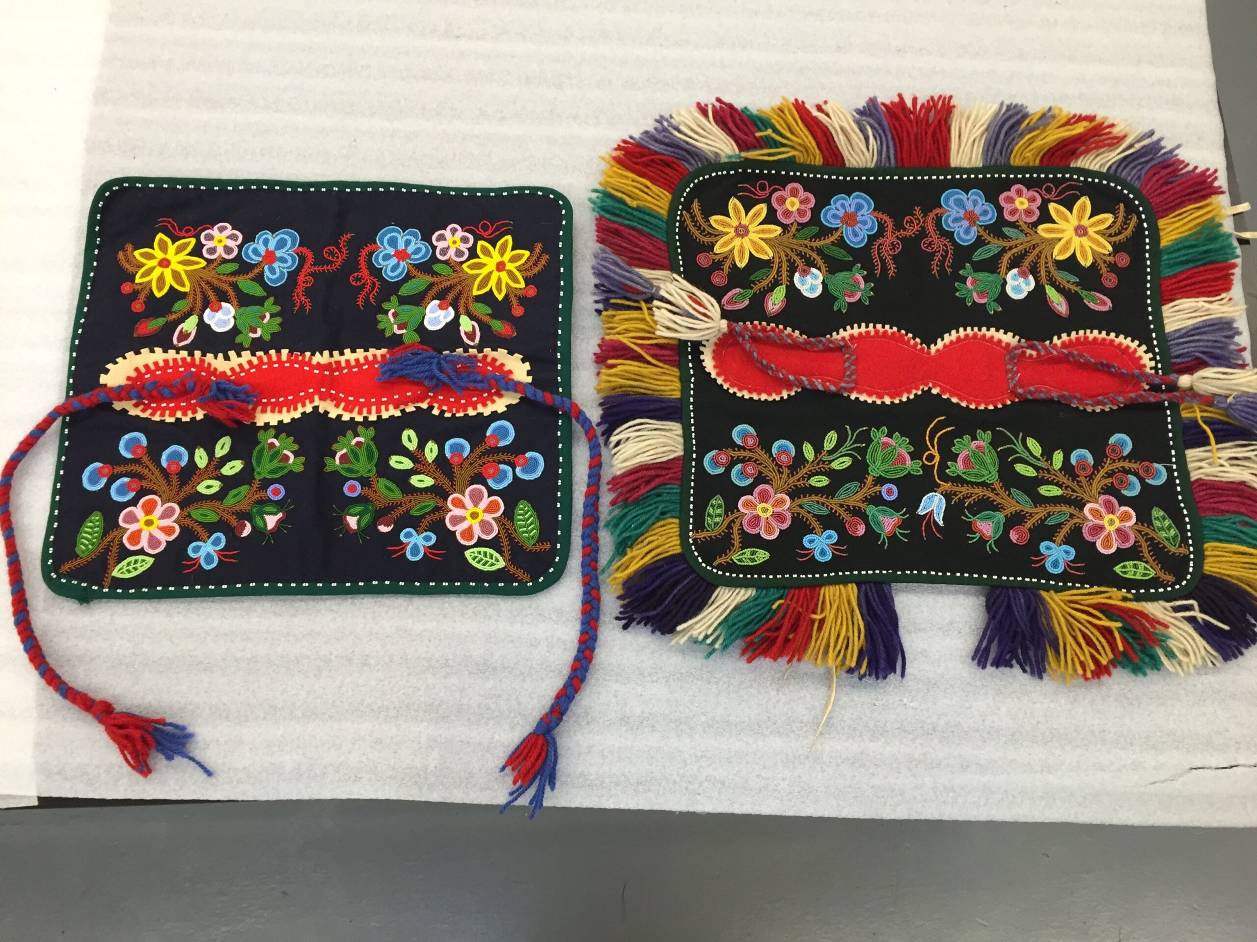 Two intricately beaded square dog blankets. The one on the right has a colourful fringe around the edges, and the one of the left does not, instead having two longer braids as ties. The pattern on the body of each piece is nearly identical.