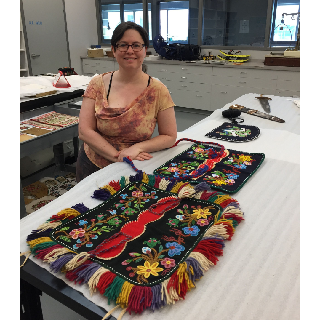 A smiling woman, Monique Olivier, standing behind a table in a Museum lab with two beaded dog blankets and a bead tea cozy laid out in front of her.