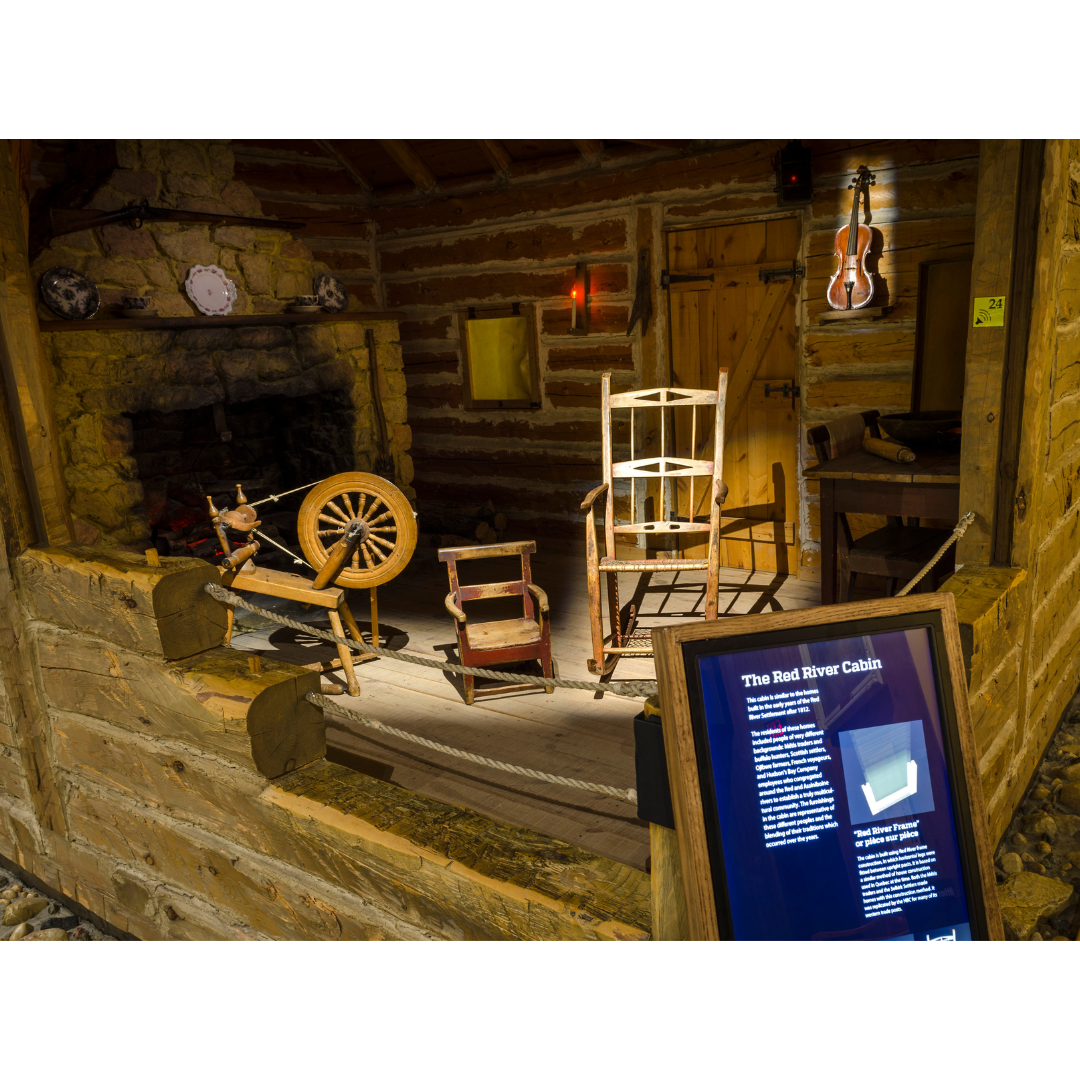 Closer view into the interior of the cabin exhibit in the Grasslands Gallery. In front of a fireplace artifacts can be seen in the room such as a spinnin wheel, a small chair, a rocking chair, and a violin, which is mounted on the far wall. In the foreground part of a digital screen is visible, where visitors can learn more about the artifacts.