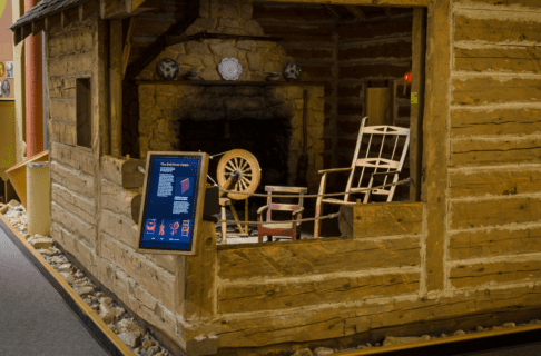 A log cabin in the Grasslands Gallery, with the close corner "cut out" to reveal inside the cabin. Inside is strategicly lit to show a fire place, rocking chair, spinning wheel, and more. At the exterior of the opened corner a digital display is mounted for visitors to engage with.