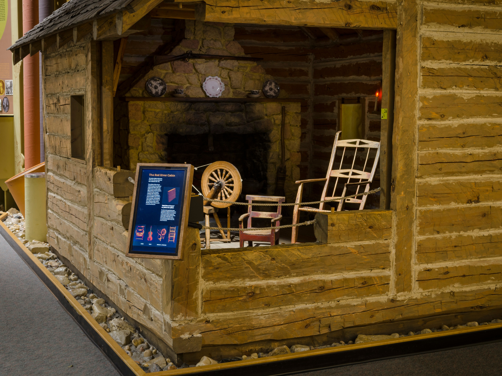 A log cabin in the Grasslands Gallery, with the close corner "cut out" to reveal inside the cabin. Inside is strategicly lit to show a fire place, rocking chair, spinning wheel, and more. At the exterior of the opened corner a digital display is mounted for visitors to engage with.