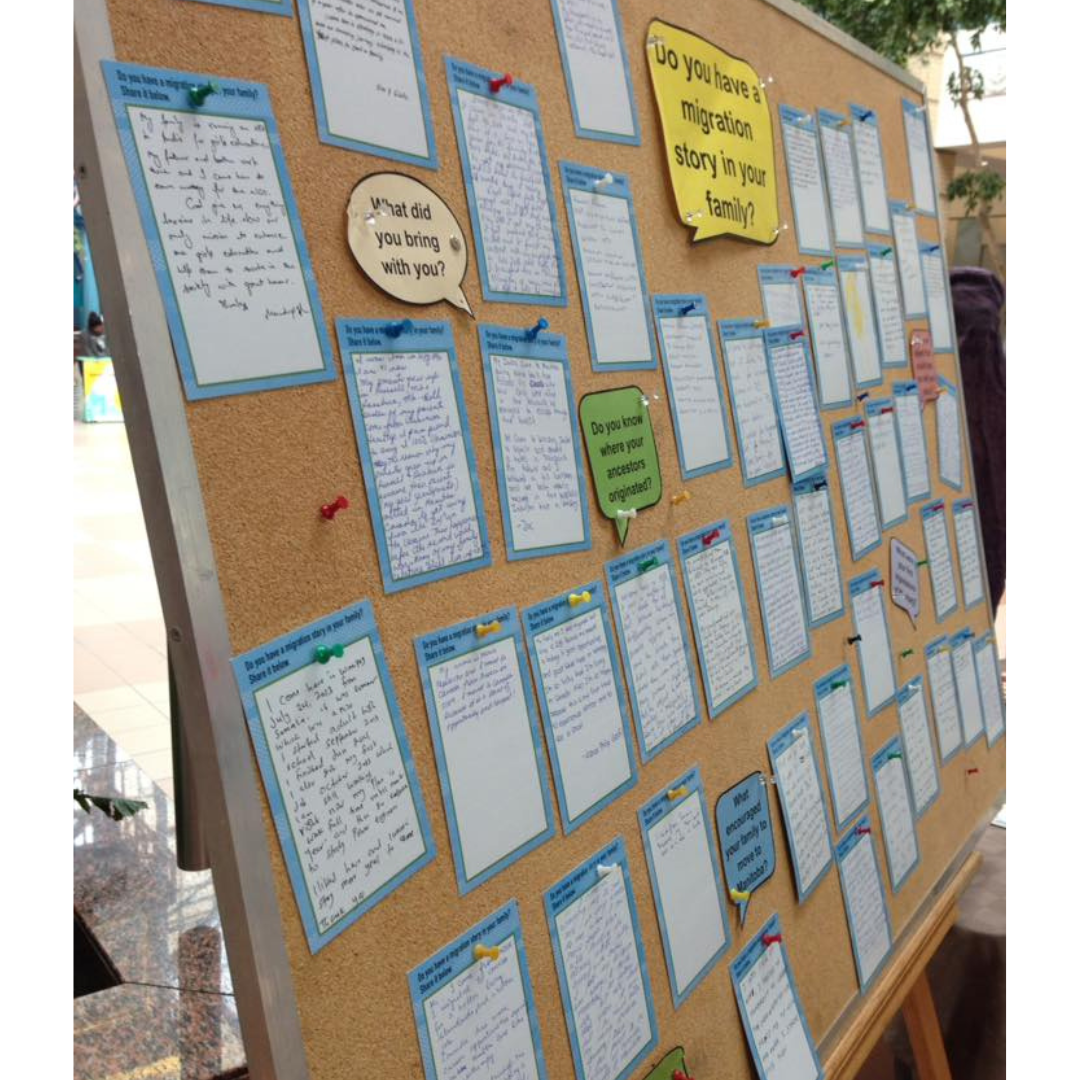 A bulletin board on an easel with several dozen notes pinned on it sharing stories and answering questions like "Do you have a migration story in your family?", "Do you know where your ancestors originated?", and "What did you bring with you?".