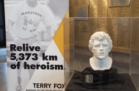 A plaster bust of Terry Fox by Leo Mol in a glass display case next to a pop up banner for the Terry Fox exhibit at the Manitoba Museum. Text on the banner reads, "Relive 5,373 km of heroism."