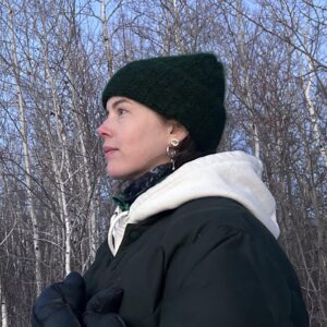 An informal headshot of Brianna Oversby in profile, standing outdoors wearing a winter coat and toque in front of birch trees bare of leaves.