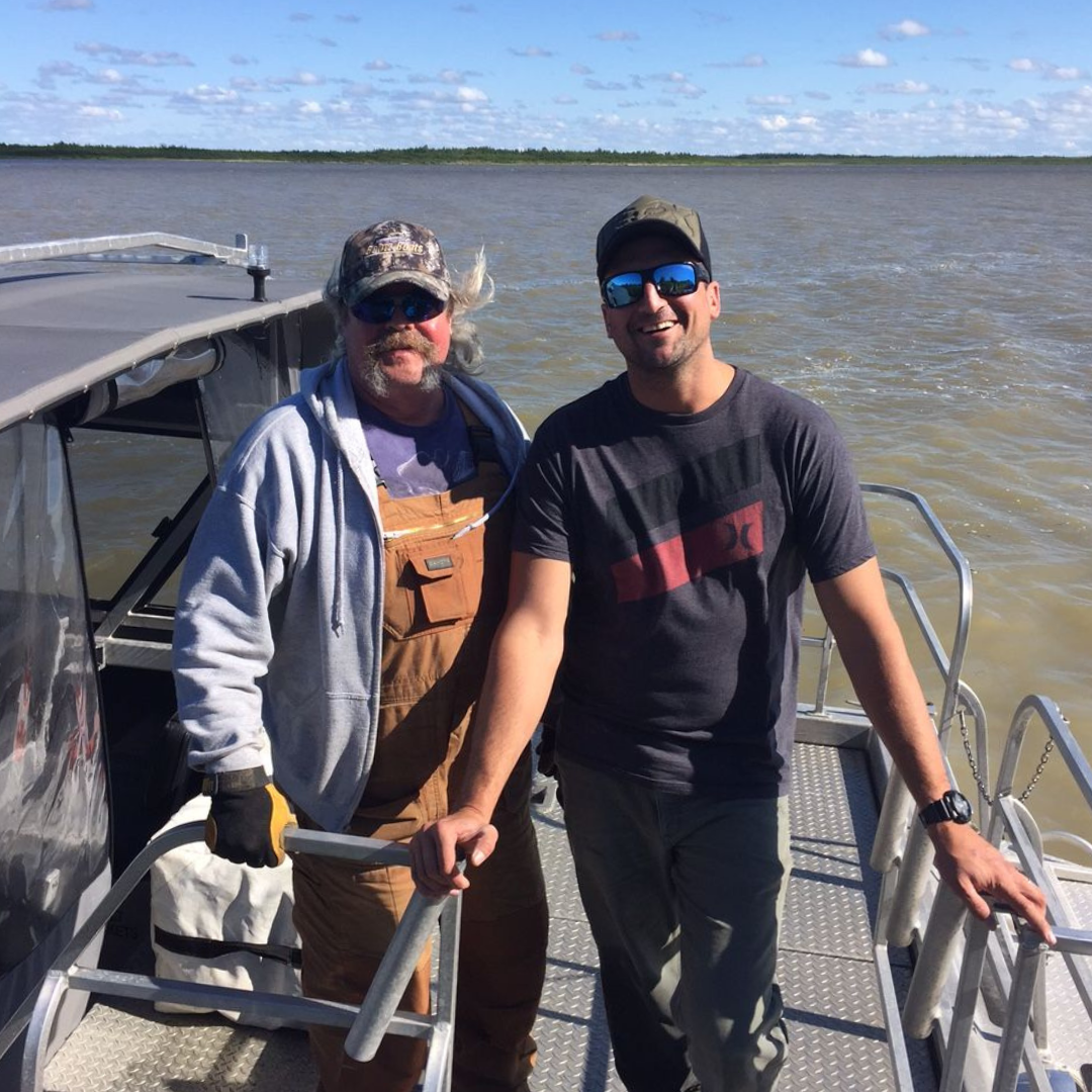 Two smiling individuals, both wearing baseball caps and sunglasses, posing on the deck of a boat for the photo.