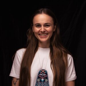 A smiling young adult with long brown hair wearing a white t-shirt and beaded octopus bag necklace.