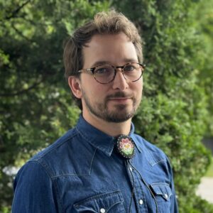 Headshot of a young adult wearing glasses, and a denim shirt with a beaded necktie.