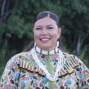 Headshot of a smiling woman outdoors, wearing a colourful jingle dress, and beaded and dentalium jewelry.