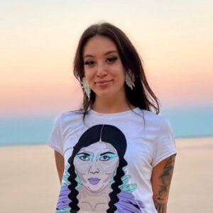 A smiling young woman posing in front of a lake in front of a dawn sky. She's wearing beaded earrings and a t-shirt with the face of a person with long, dark braids on it.