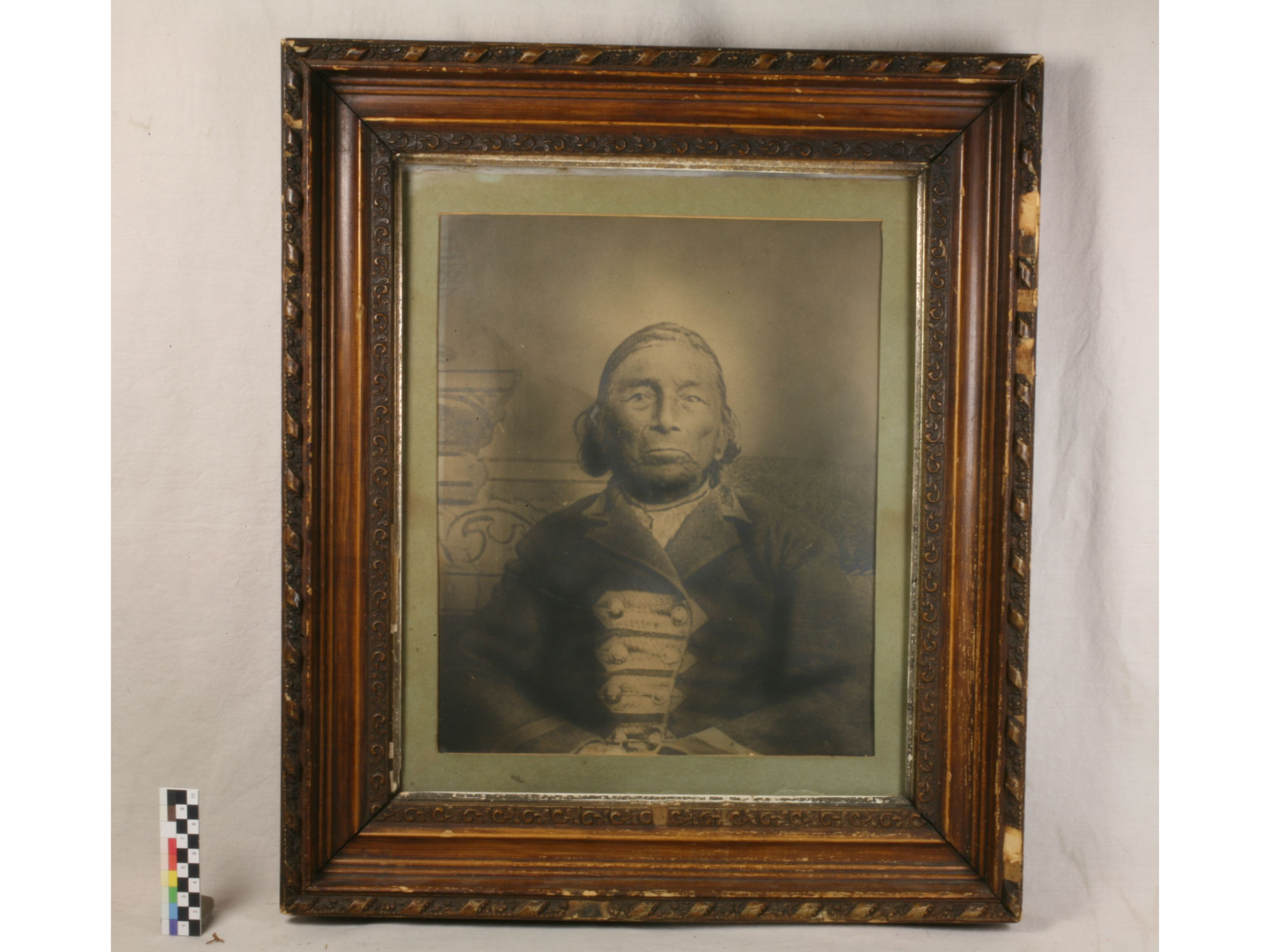 A sepia toned formal photograph of a serious-faced man, seated against a faint backdrop and where a military-style coat. The photograph is in a ornate wooden frame. Both frame and photograph look aged, and a little worn and torn. The matte around the photograph has aged to a greenish colour.