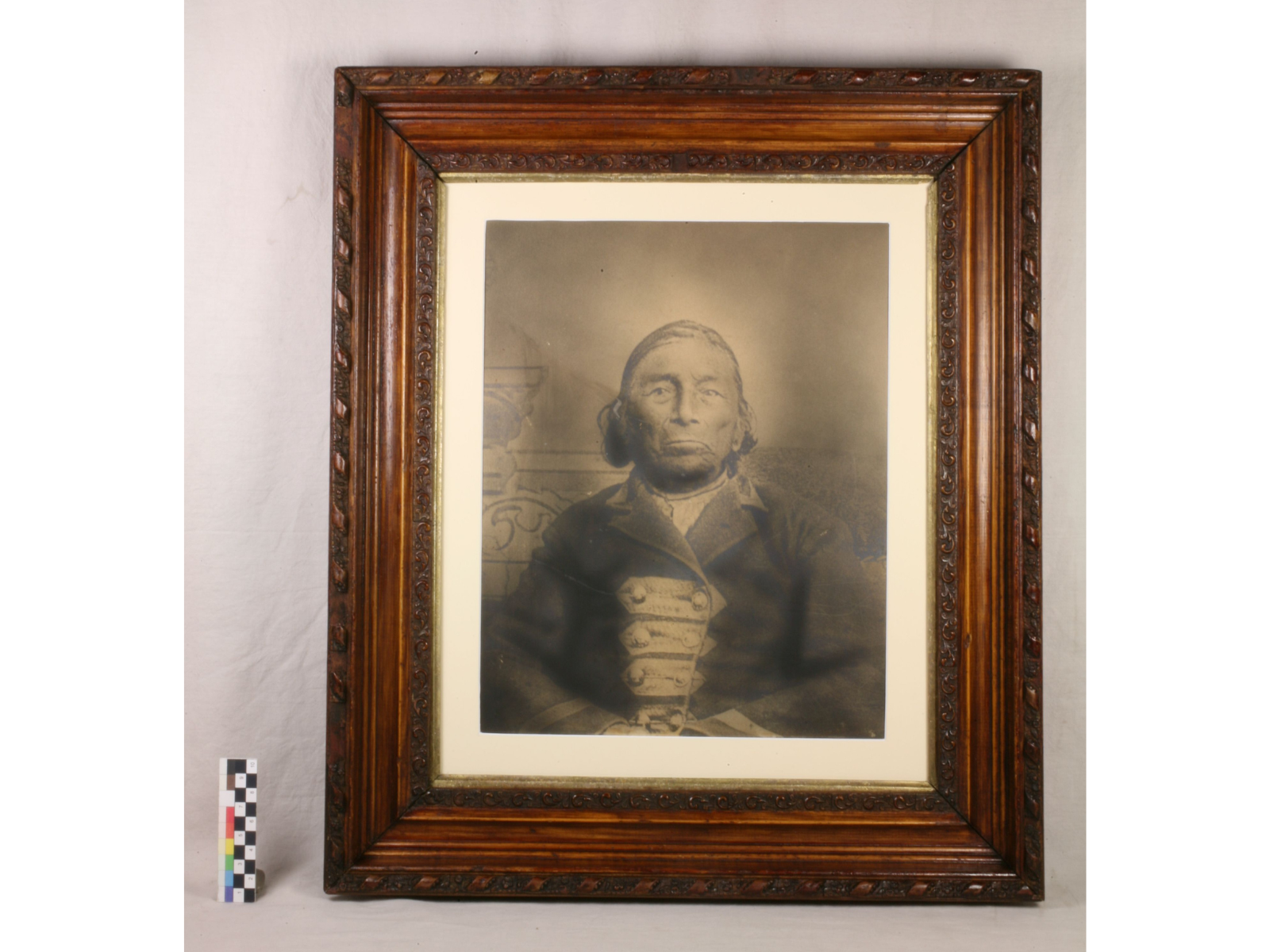 A sepia toned formal photograph of a serious-faced man, seated against a faint backdrop and where a military-style coat. The photograph is in a ornate wooden frame. The frame and glass have been cleaned, and the matte replaced with a fresh white one, making the features and contrasts of the image clearer.