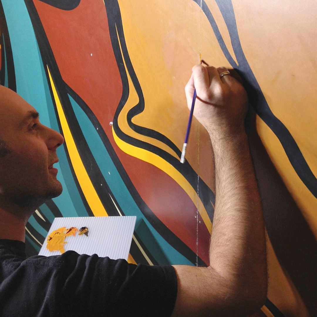 Close-up from behind and to the side on a person holding a paint palette and a paintbrush, touching up a orange portion of the mural.