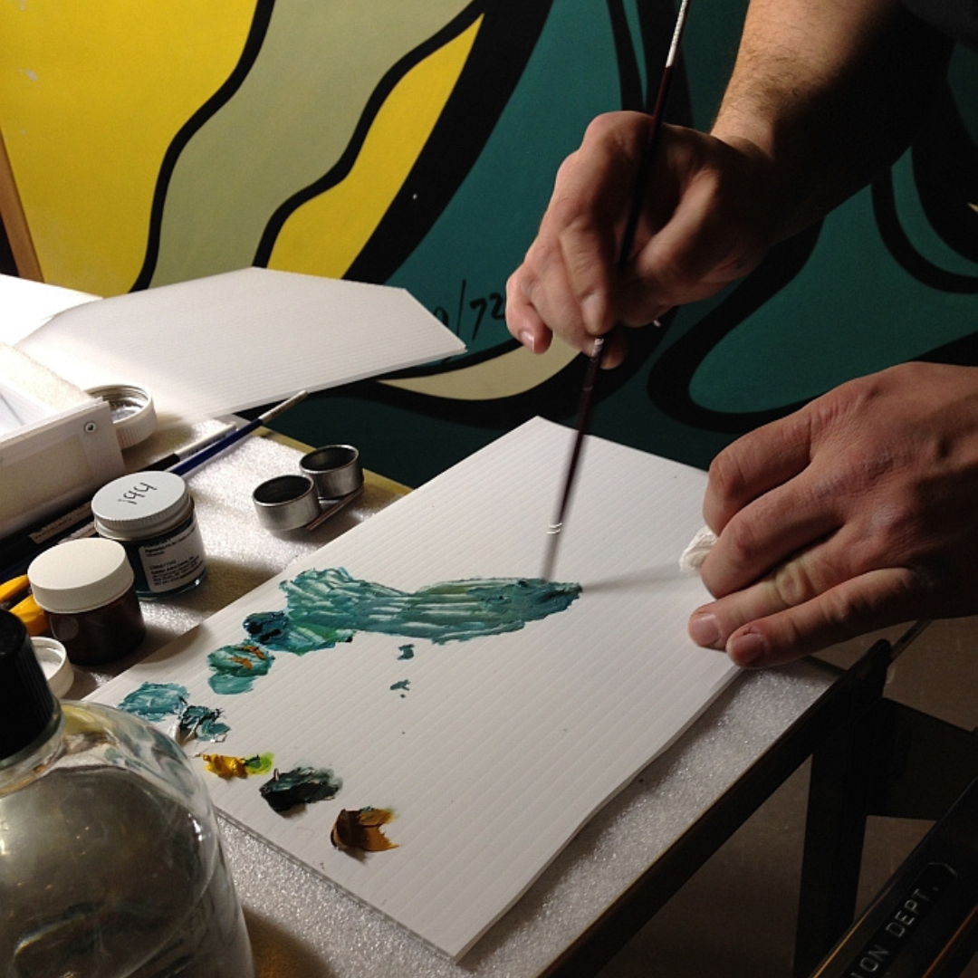 Close up on a person's hands as they mix a custom teal blue-green colour of paint on a board.