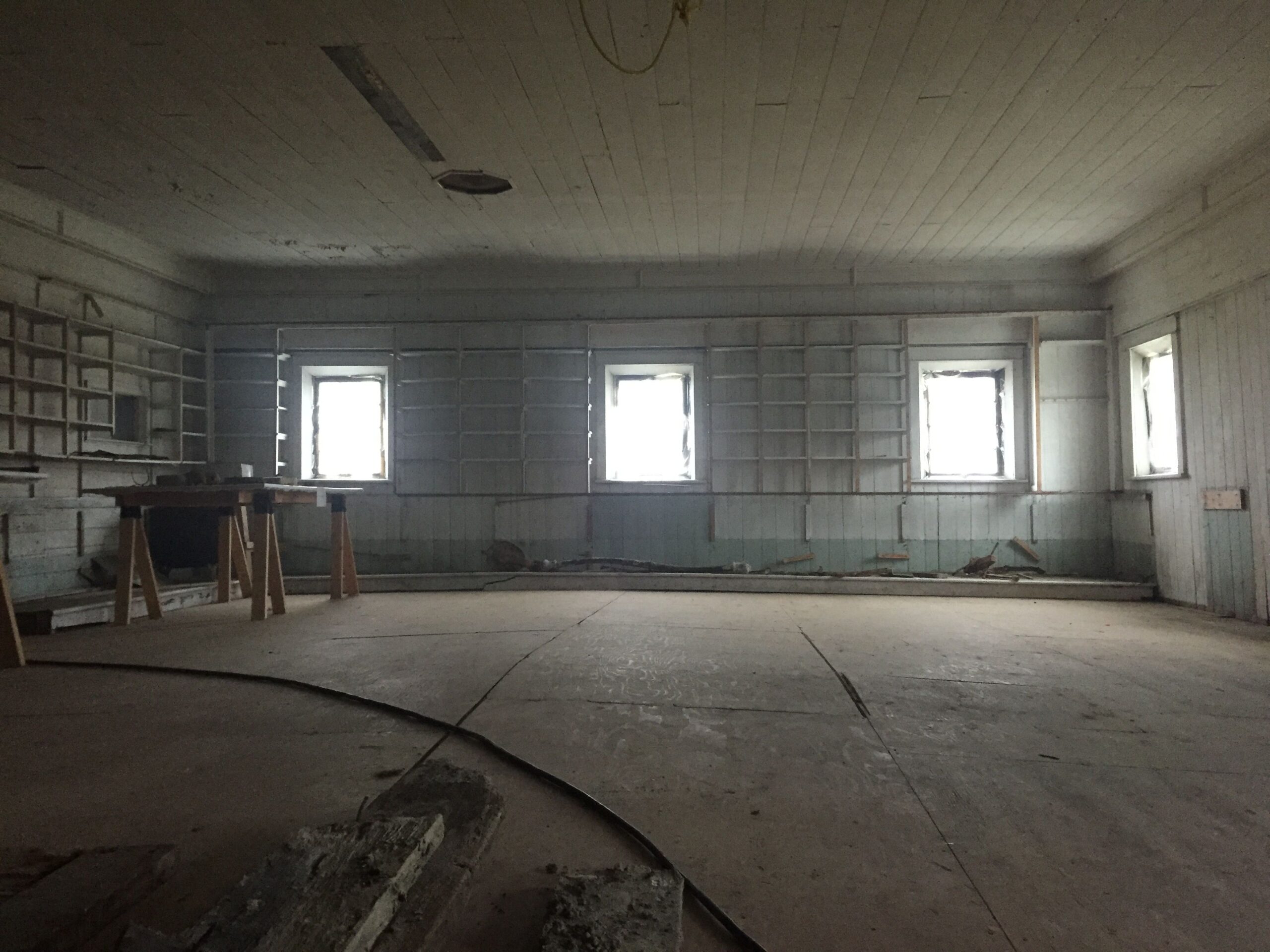 An empty room, lit only by the sunlight coming through four windows. Along the walls are empty build-in shelves, and to the left is a wide board laid across two saw horses like a table.