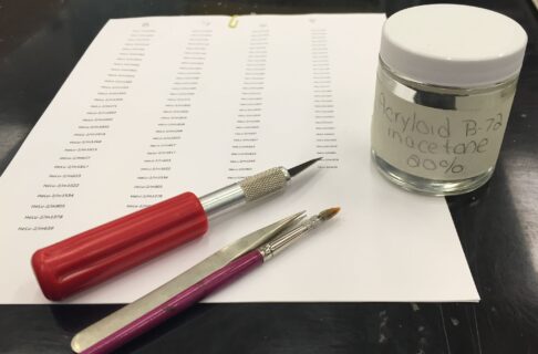 A sheet of paper containing columns of catalogue numbers, with a cutting tool, tweezers, small paintbrush, and a jar of sealant placed on top.