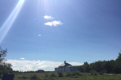 View across a stretch of grass and bush towards a large three-storeyed building beneath a blue sky with occasional white clouds.