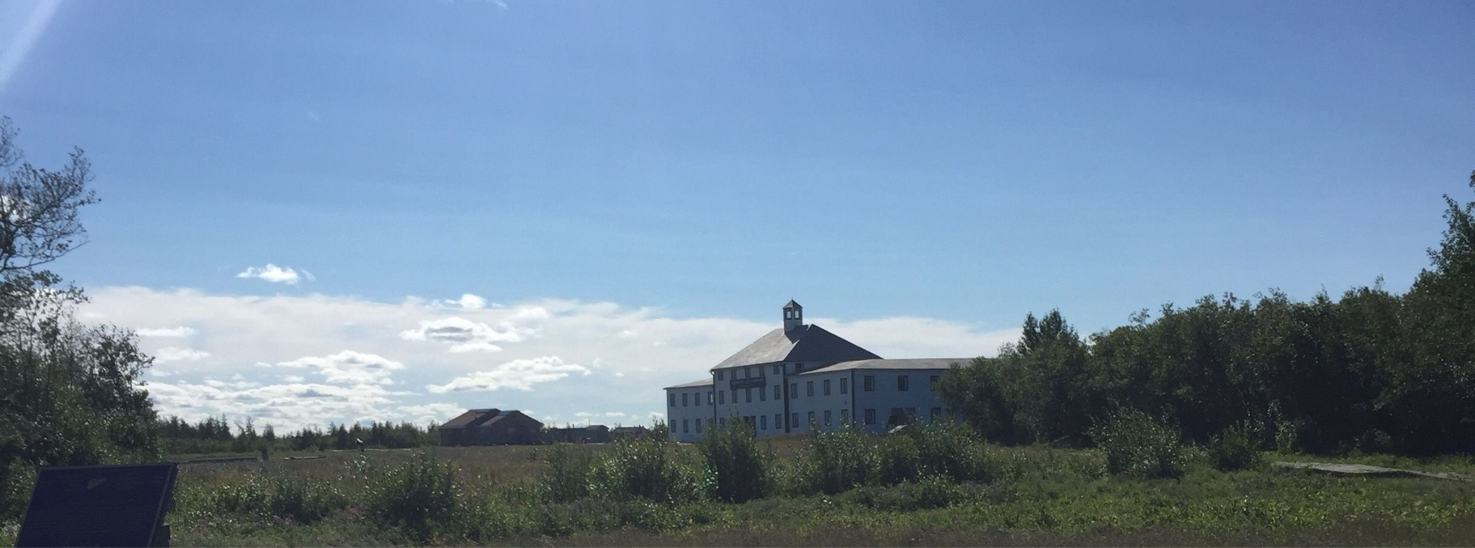 View across a stretch of grass and bush towards a large three-storeyed building beneath a blue sky with occasional white clouds.