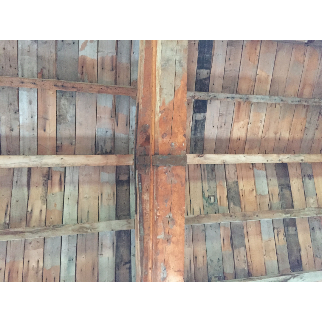 Looking directly up at a centre peak of a wooden ceiling, the wood planks have slight variations in colour from dark greys to light browns and reds.