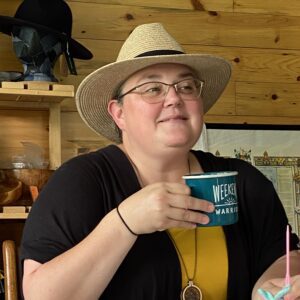 Informal headshot of Melanie Gamache sitting in a wooden walled room, wearing a wide brimmed hat, and holding up a teal coloured mug.