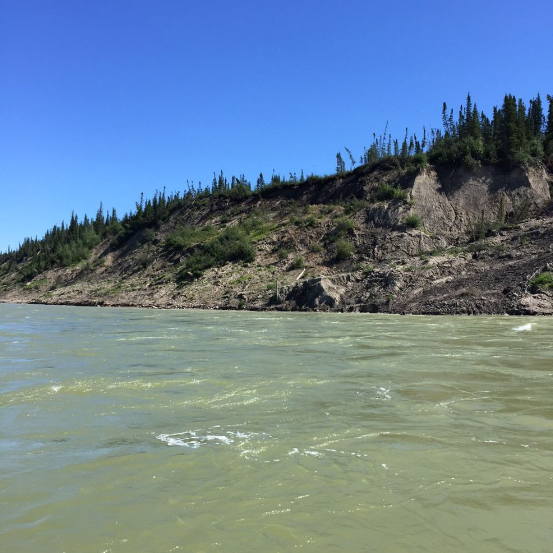 A rocky, treed bank increasing in incline, along a cloudy river.