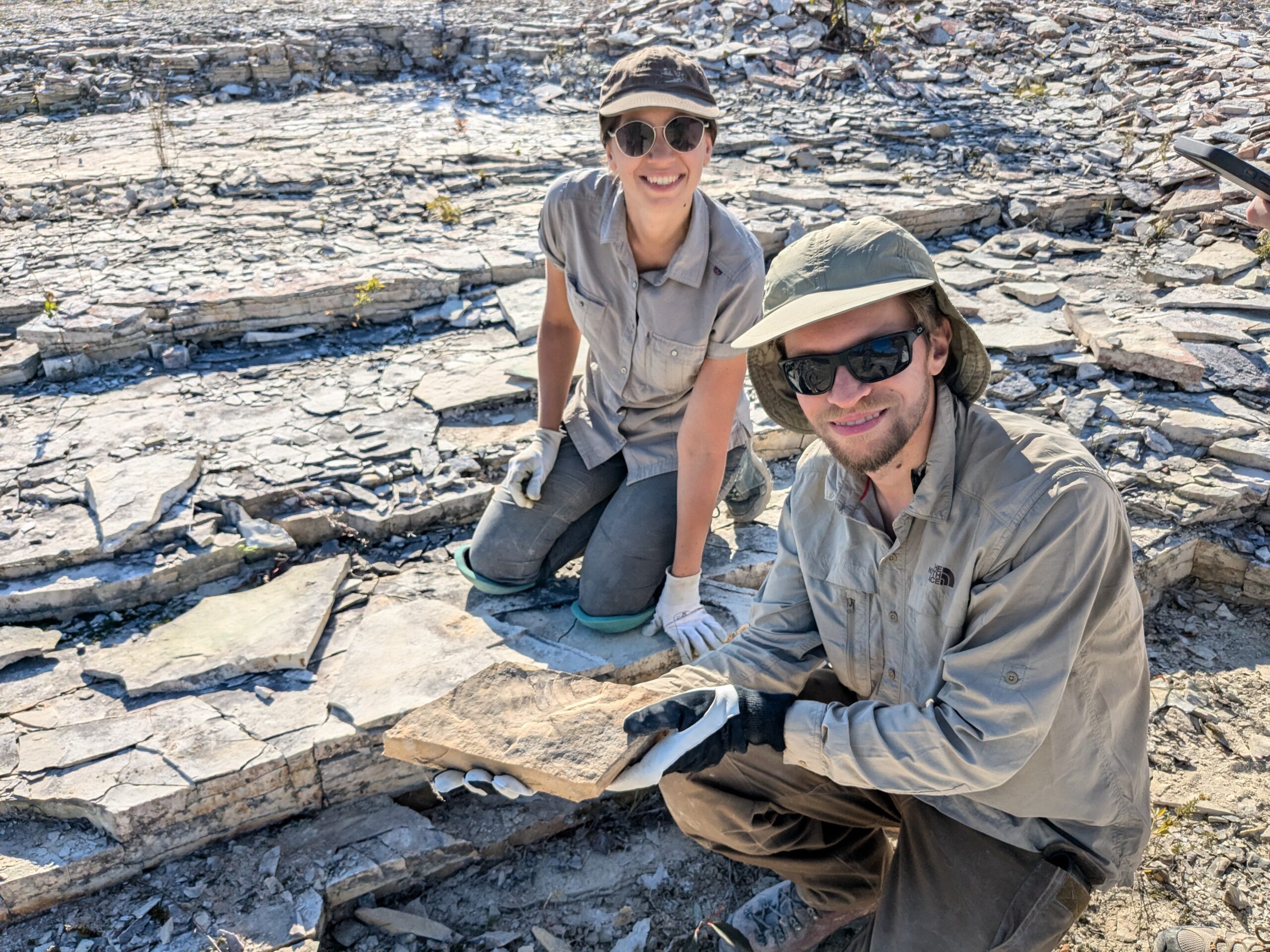 Dr. Joe Moysuik and a coleague on a rocky outcropping holding up a recently found fossil specimen.