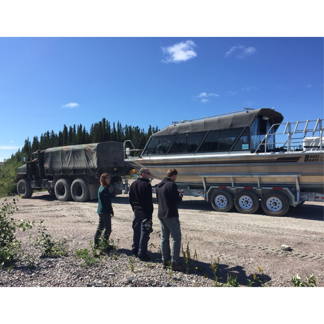 Three individuals standing to the side of a dirt road, on which a large truck is pulling a trailer with a motor boat on it.