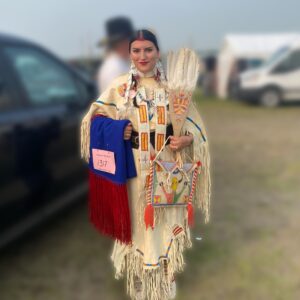 A young woman wearing a traditional Anishinaabe dress holding up a beaded blanket, bandolier bag, and fan.