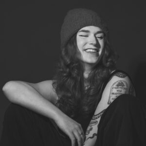 Seated black and white headshot of a smiling young woman with her arms rested on her raised knees. Several tattoos adorn her left arm, and she's wearing a beanie.