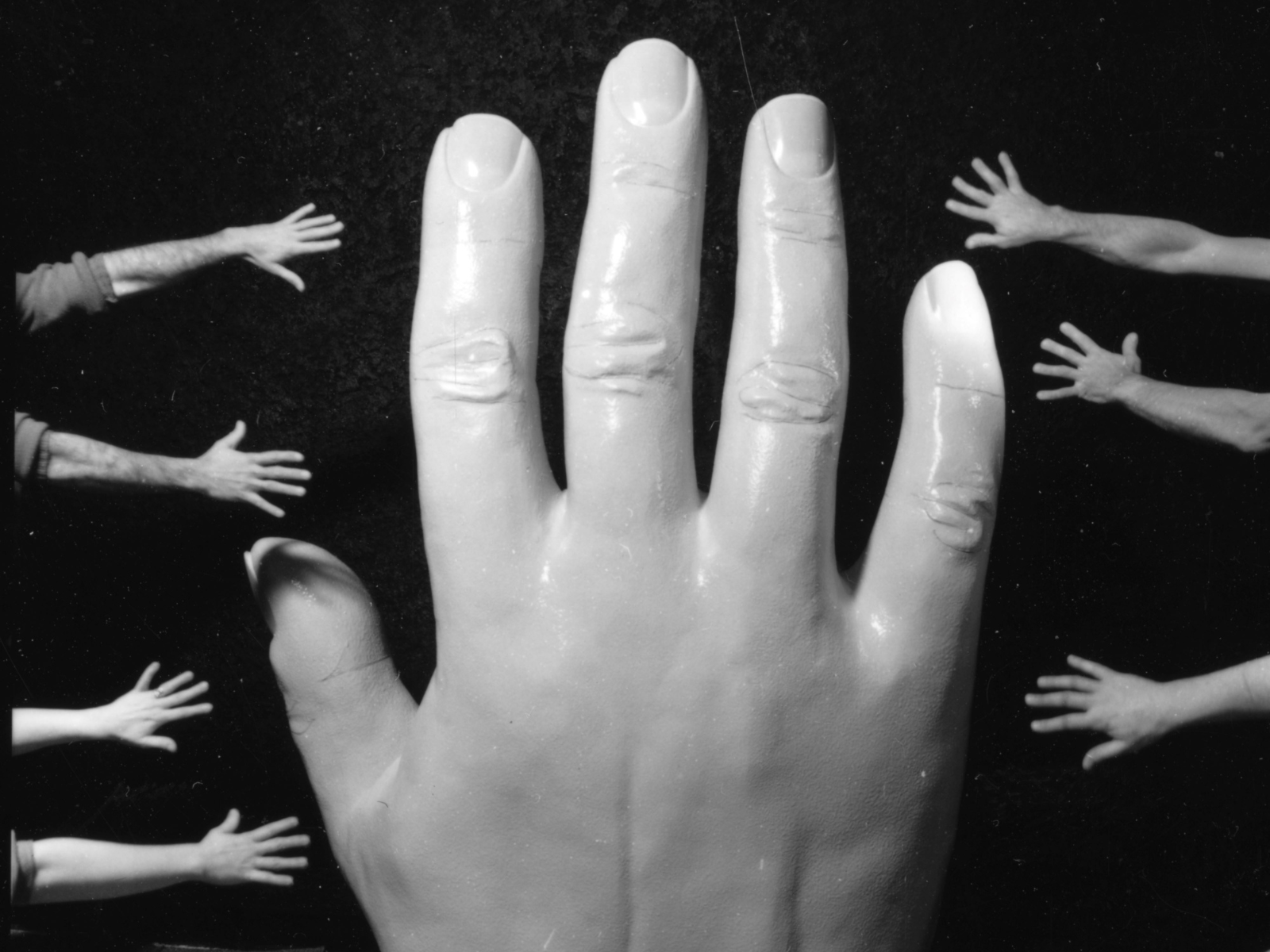 A black and white photograph of a giant hand from the bottom of the frame, with seven hands reaching into frame from the sides.