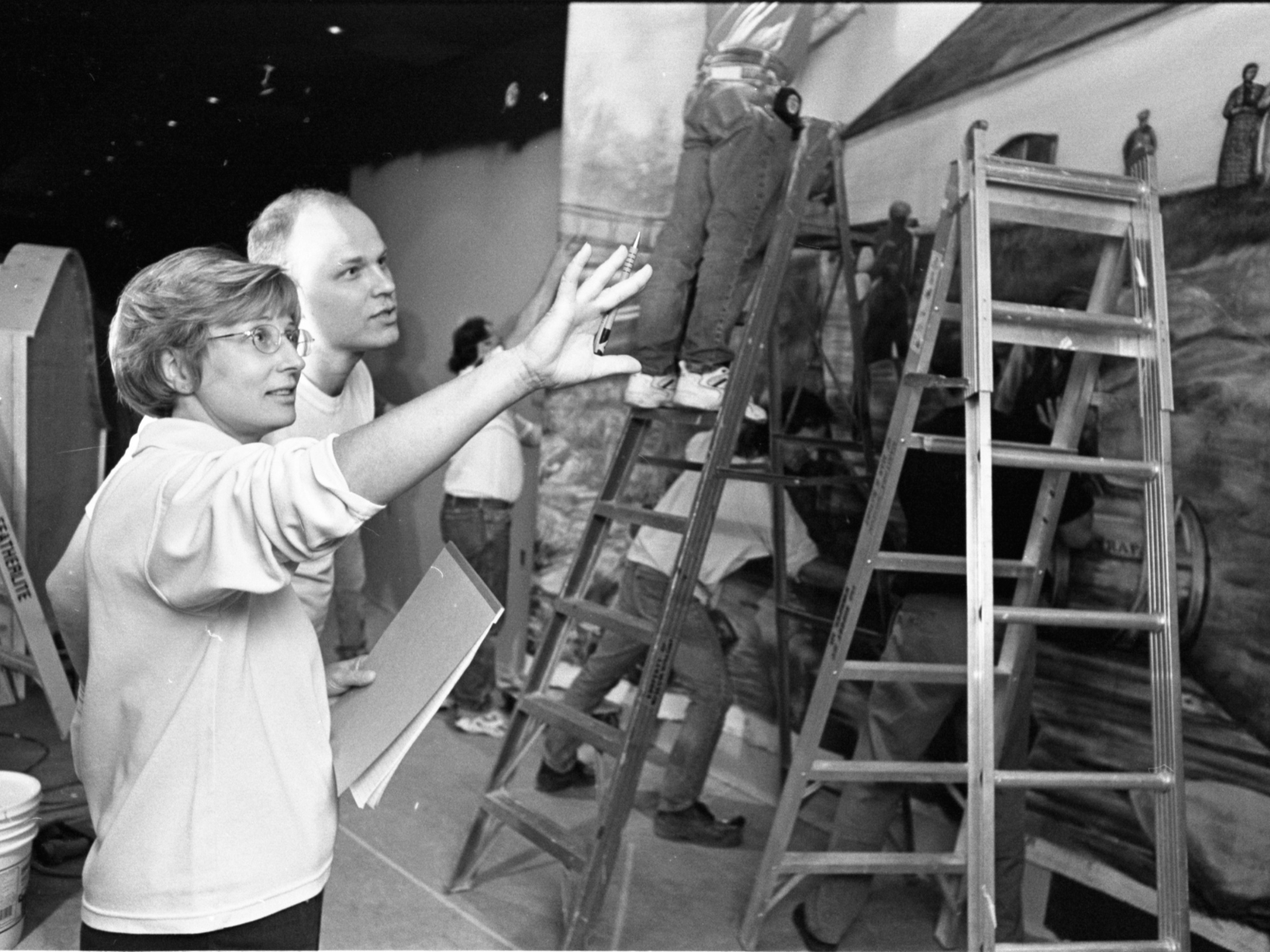 Two individuals in discussion during construction of a hall exhibit. Several ladders are in place along the wall to facilitate construction.