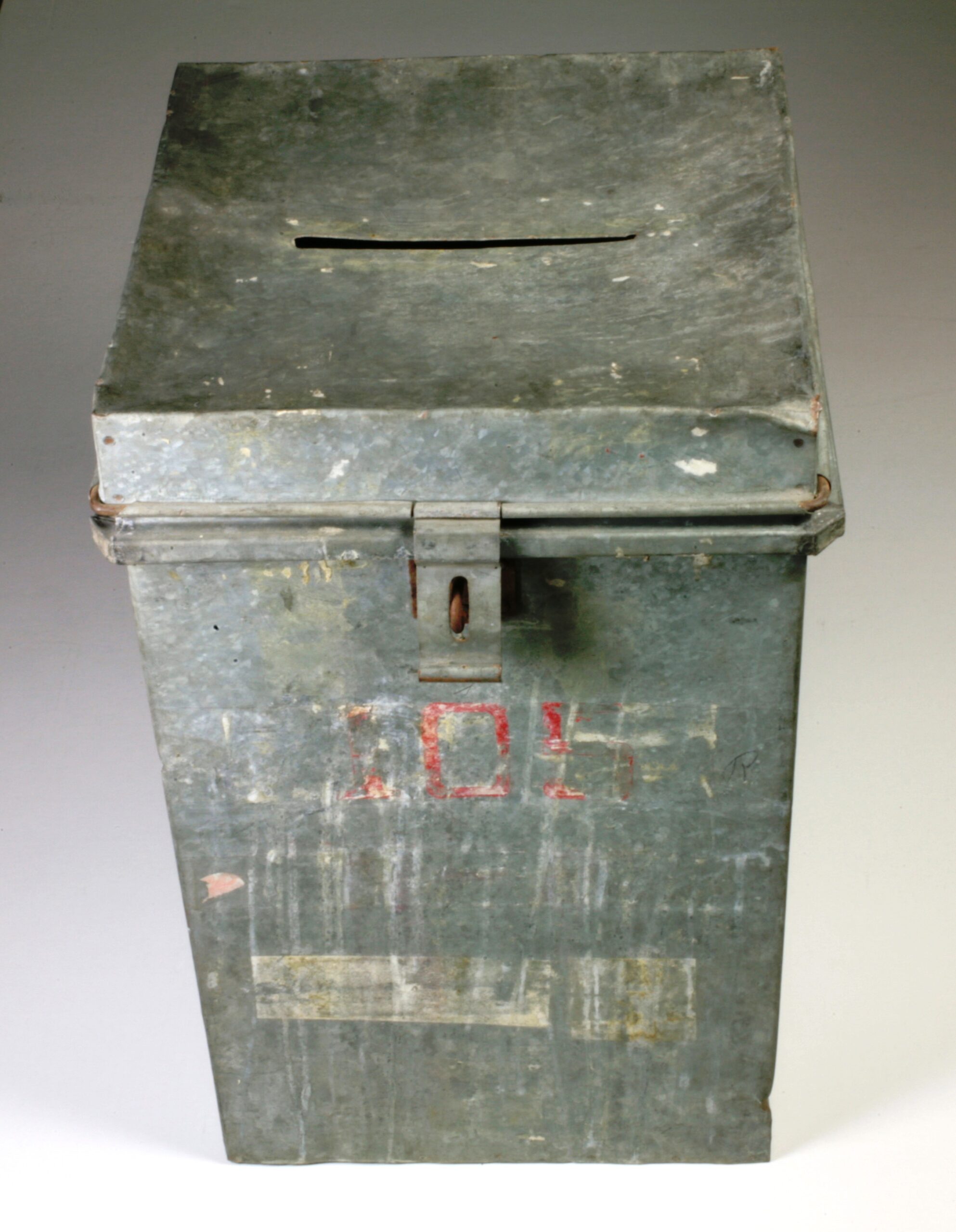 An old, worn ballot box with a latch for a lock on the front and a slit for votes in the lid. Still faintly visible is the number 105 in red.