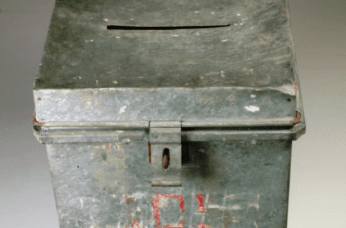 An old, worn ballot box with a latch for a lock on the front and a slit for votes in the lid. Still faintly visible is the number 105 in red.