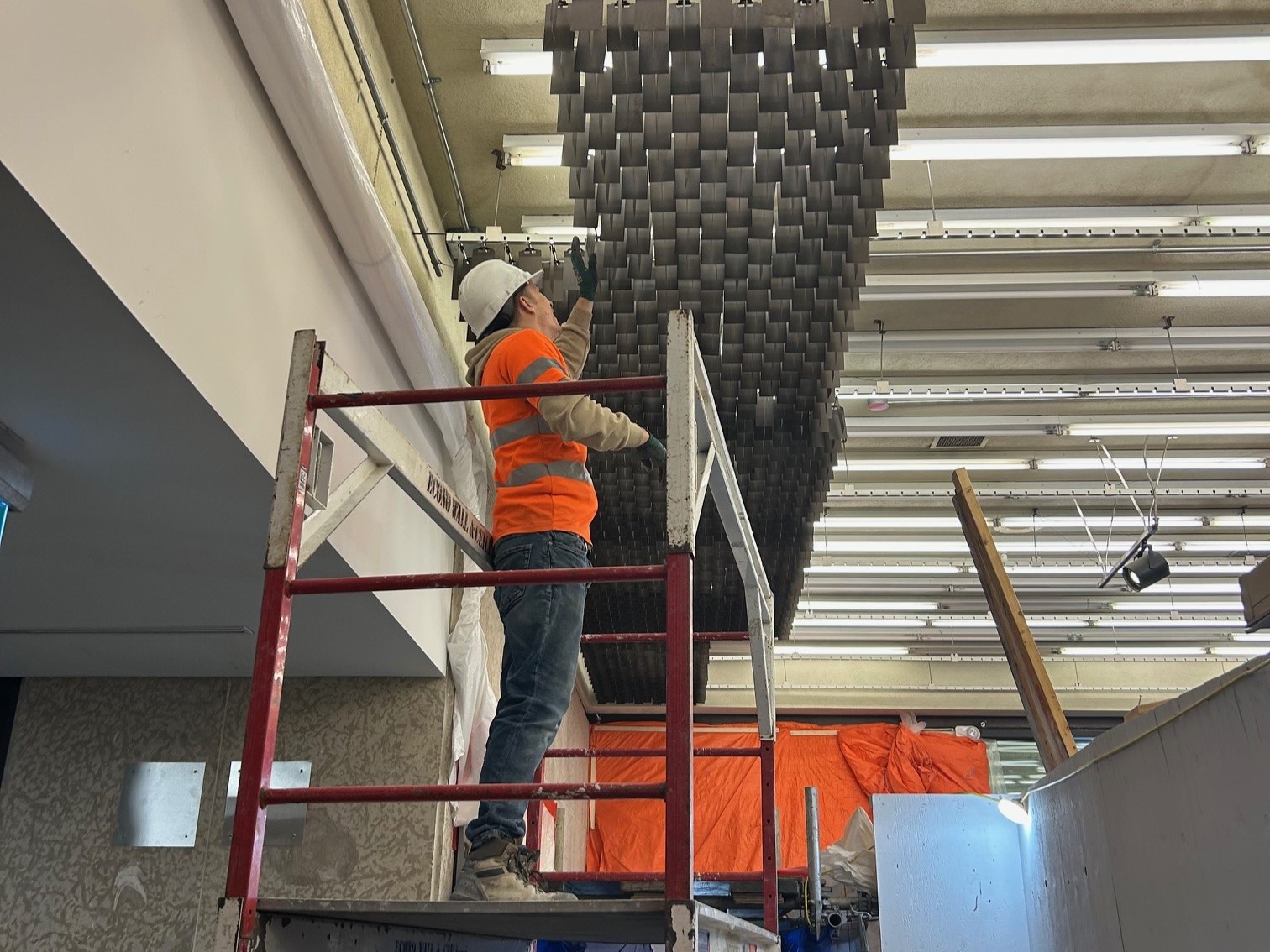 A construction worker in a high vis shirt and hard hat stands on a platform, reaching up to remove a panel of the ceiling tiles in the Museum Foyer.