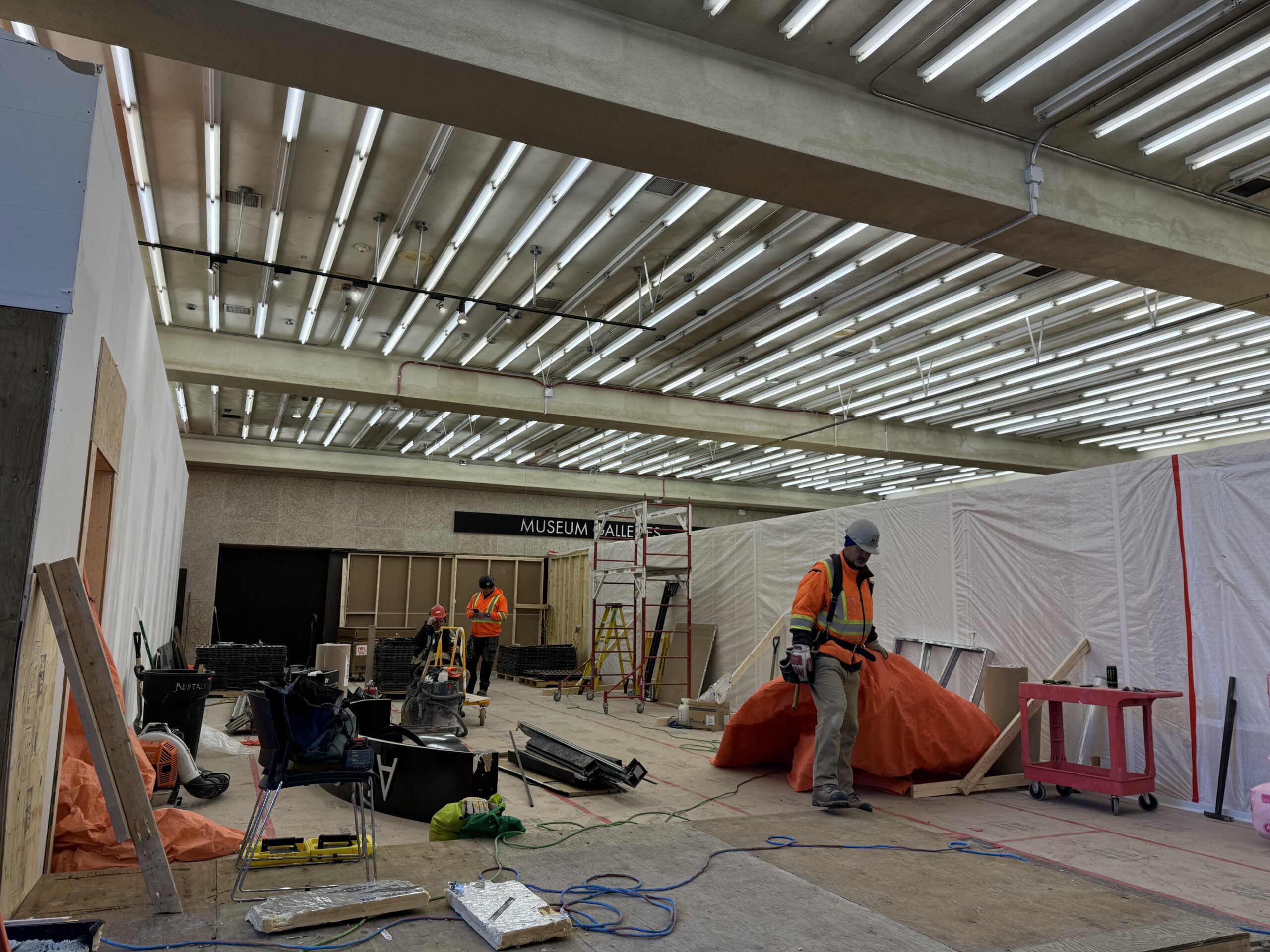 Inside the hoarded off section of the Museum foyer which is now a constrcution site. The ceiling tailes have been removed, exposing the base of the ceiling. Workers in high-vis and hard hats move about the space.