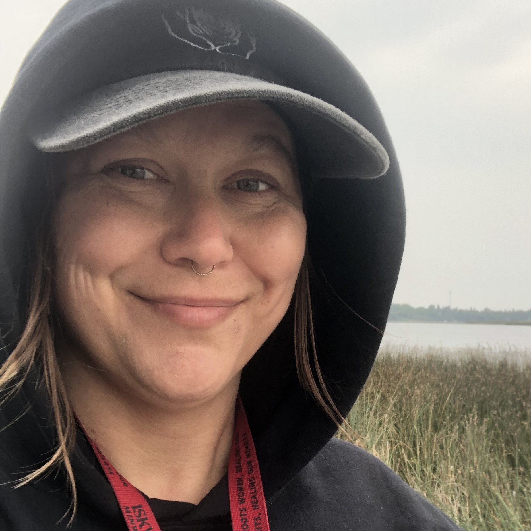 Selfie of a smiling woman wearing a baseball hat and hoodie with the hood up standing by a wetland and body of water.