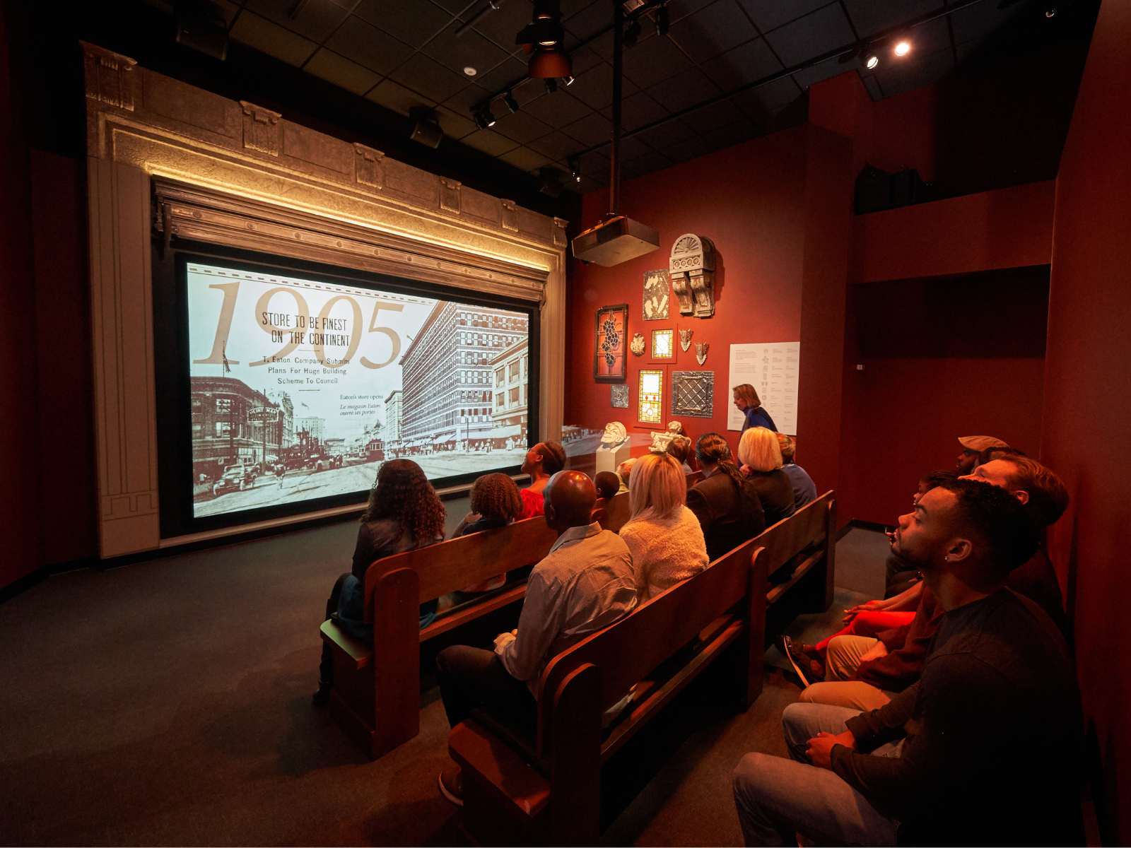 A group of people seated in pews in a small theatre, with a screen framed in ornate stonework. On the screen a video plays showing a timeline of the history of Winnipeg. On the far wall are several artifacts and architectural accents including a stone grotesque.