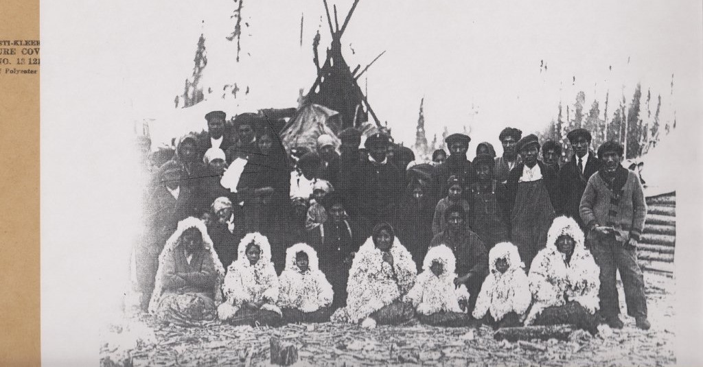 A historical black and white photograph showing a group of people, many wrapped in layers and furs, standing and sitting together for a photo in front of a tipi.