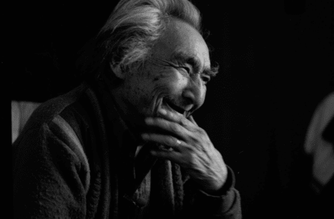 A black and white portrait of a seated older man from the side, smiling and laughing behind his hand.