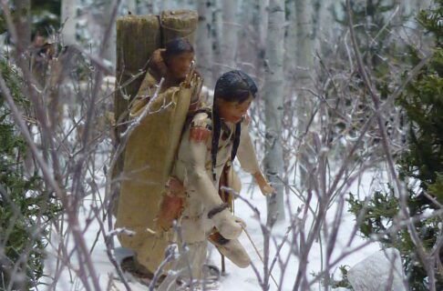 Two figures in a miniature diorama winter scene, wearing traditional winter gear and with bundles on their backs.