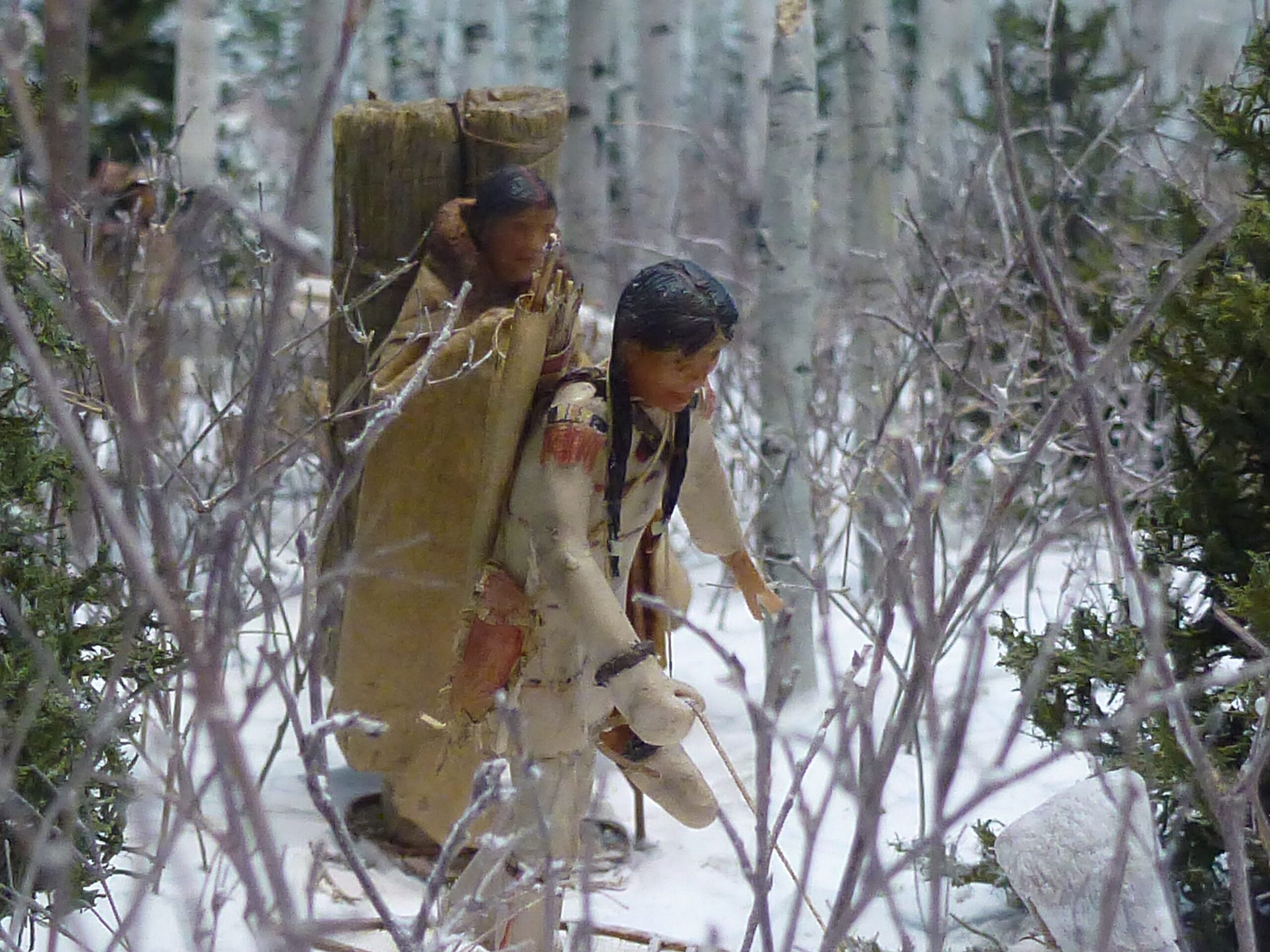 Two figures in a miniature diorama winter scene, wearing traditional winter gear and with bundles on their backs.