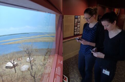 Two individuals each holding cell phones standing in front of the Aschkibokahn diorama in the Manitoba Museum's Lower Parklands Gallery.