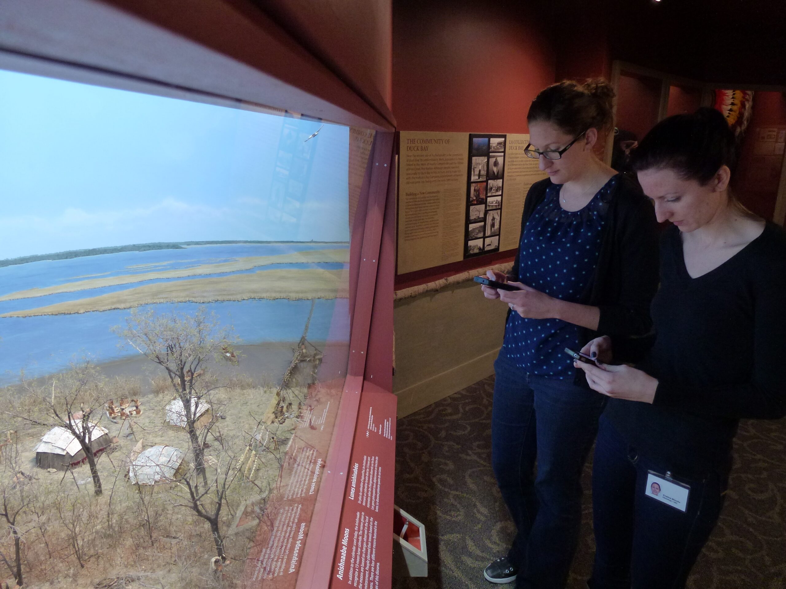 Two individuals each holding cell phones standing in front of the Aschkibokahn diorama in the Manitoba Museum's Lower Parklands Gallery.