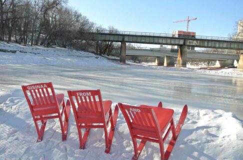 Three red chairs with skiis on their feet in a snowbank along the side of a cleared, frozen river. The backs of each chair has a word written on it in white, "Gisinaa", "From", "And".